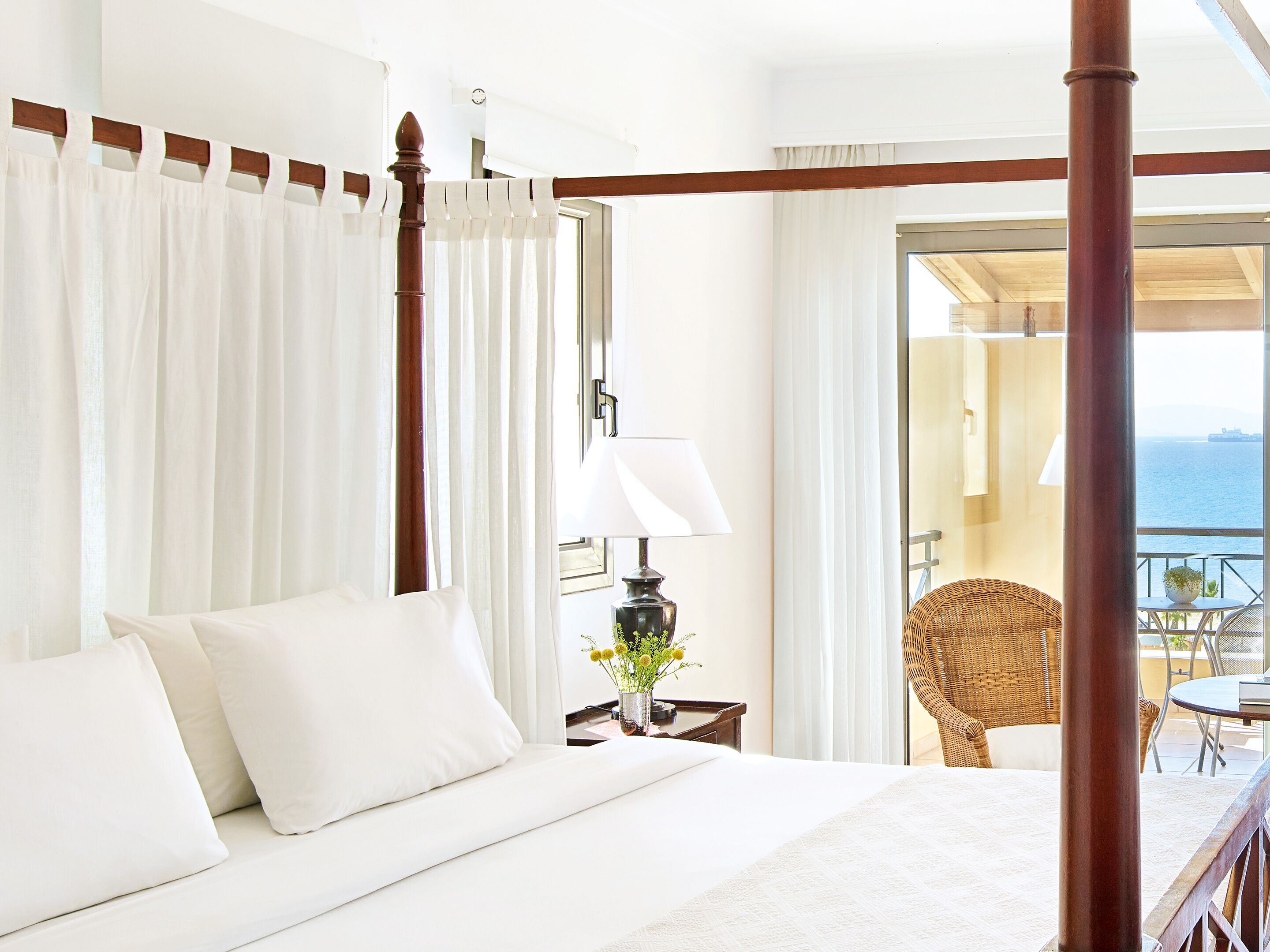 Een hemelbed met witte lakens, een nachtkastje met een lamp en een bloemenvaas, en uitzicht op zee door een open balkondeur.