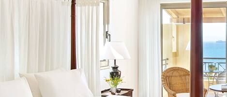 A four-poster bed with white linens, a bedside table with a lamp and a vase of flowers, and a view of the sea through an open balcony door.