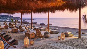 En avslappnad lounge vid stranden med parasoller av halm, solstolar och bord av trä.