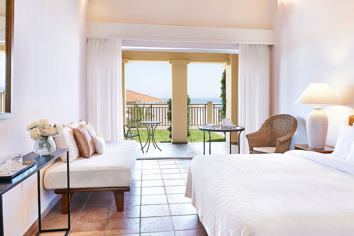 Habitación de hotel con una cama, una silla, una mesita y vista al mar a través de una puerta abierta.