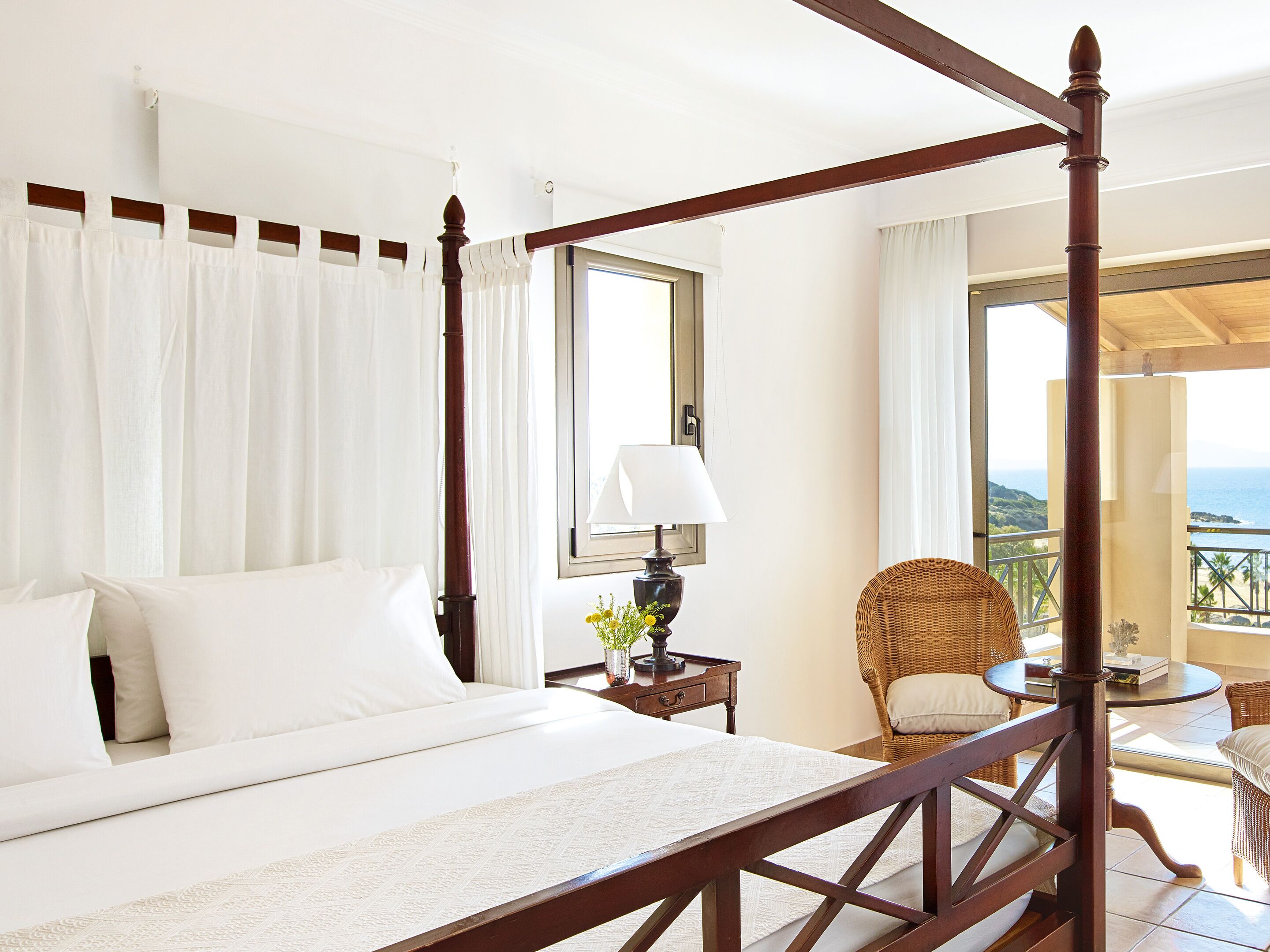 A four-poster bed with white linens, a bedside table with a lamp and flowers, a wicker chair, and a balcony with a view of the ocean.