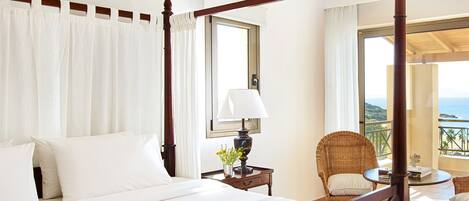 A four-poster bed with white linens, a bedside table with a lamp and flowers, a wicker chair, and a balcony with a view of the ocean.