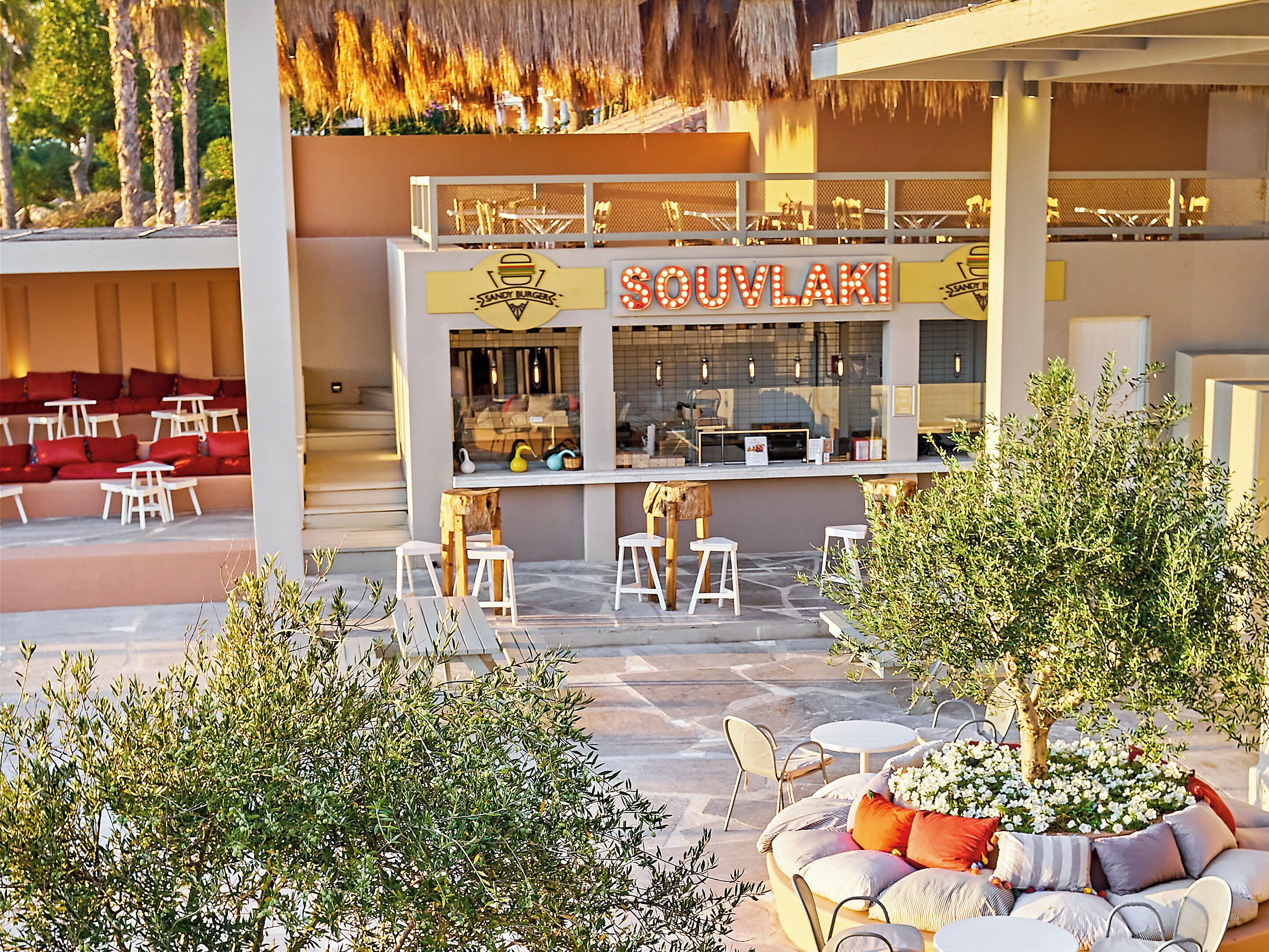 Een buitenruimte met een bar, genaamd "SOUVLAKI", en een pool lounge met rode kussens.