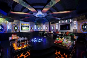 Zona de bar con varias pantallas, una barra llena de bebidas y paneles decorativos en la pared.