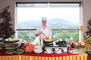 Montaje de bufé con un chef preparando alimentos, vista de un paisaje montañoso y diversos platos y utensilios sobre la mesa.