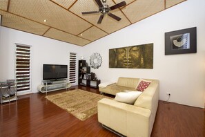 A living room with a ceiling fan, a flat-screen TV, a beige sofa, a coffee table, and a large abstract painting. - Serene Place (Nelly Bay)