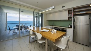 Een moderne keuken met een houten tafel en witte stoelen, een roestvrijstalen koelkast, en uitzicht op zee door grote glazen deuren.