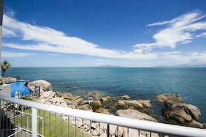 Ein Poolbereich mit klarem, blauem Wasser, umgeben von großen Felsen und Blick auf das Meer sowie entfernte Berge.
