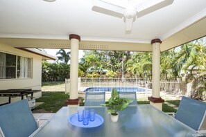 An outdoor patio with a glass table, chairs, a ceiling fan, and a view of a swimming pool and greenery. - Island Dreams (Nelly Bay)