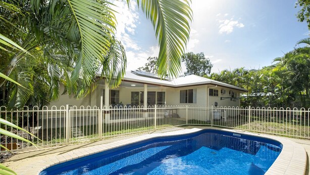 A swimming pool with a white fence, surrounded by palm trees and a house with a covered patio. - Island Dreams (Nelly Bay)