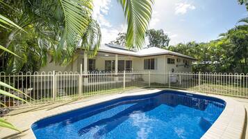 A swimming pool with a white fence, surrounded by palm trees and a house with a covered patio.