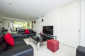 A modern living room with a black leather sofa, a red cushion, a glass coffee table, and a flat-screen TV.