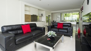 A modern living room with two black leather sofas, a glass coffee table, and a kitchen in the background.