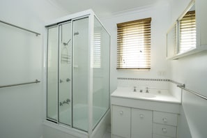 A modern bathroom with a glass shower enclosure, a white sink with a mirror, and a window with blinds.
