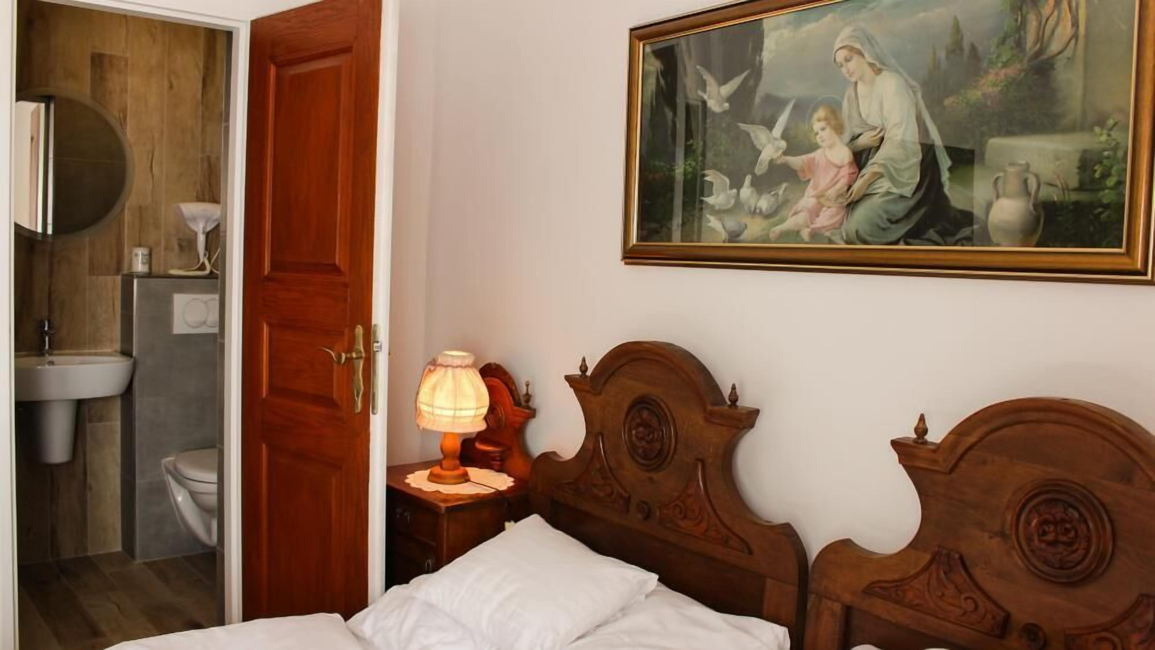 Une chambre à coucher avec une tête de lit en bois, une table de chevet avec une lampe, un tableau représentant une femme et un enfant, et une salle de bain visible à travers une porte ouverte.