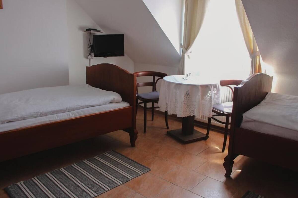 A small, well-lit room with two wooden beds, a single chair, a small table with a lace tablecloth, and a television mounted on the wall.