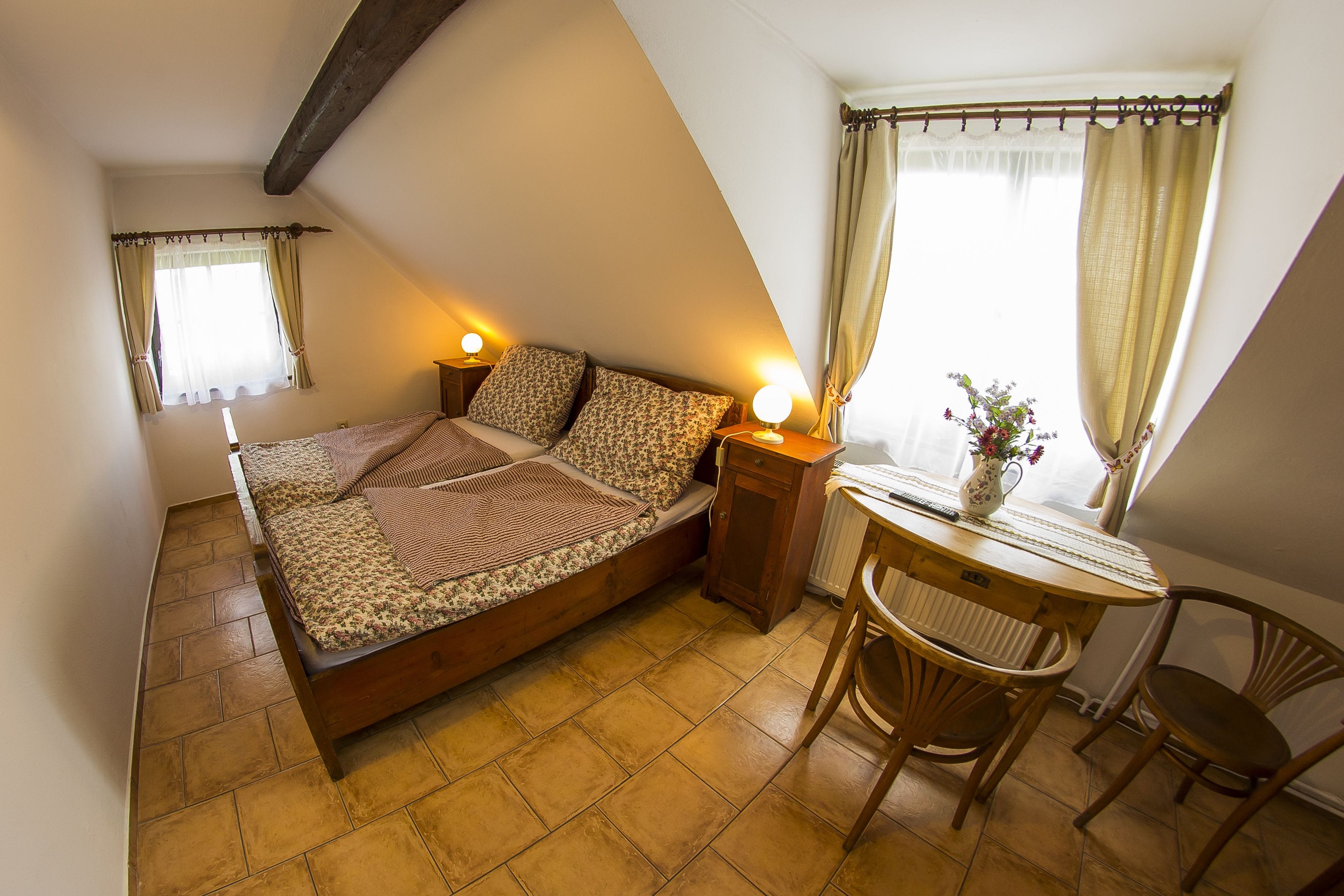 Un dormitorio con cama, lámparas de noche, una mesita con una silla y una ventana con cortinas.