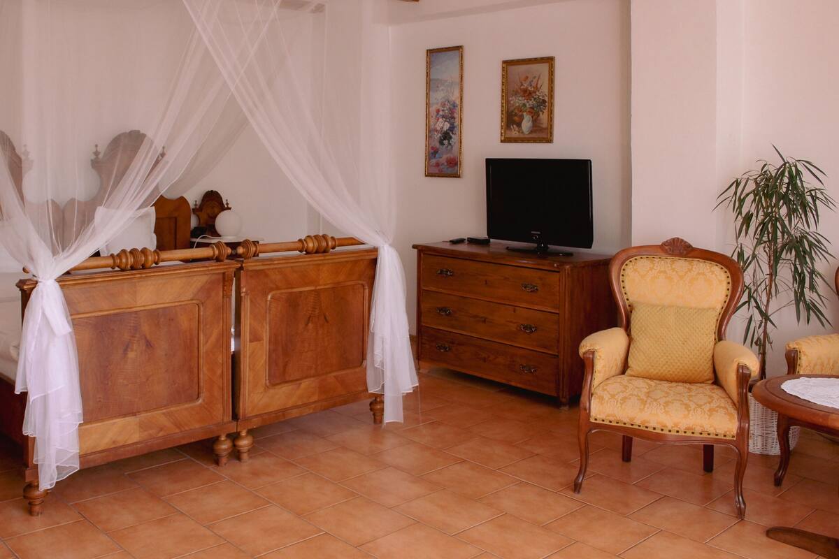 A bedroom with a four-poster bed, a wooden dresser, a television, a chair, and a potted plant.