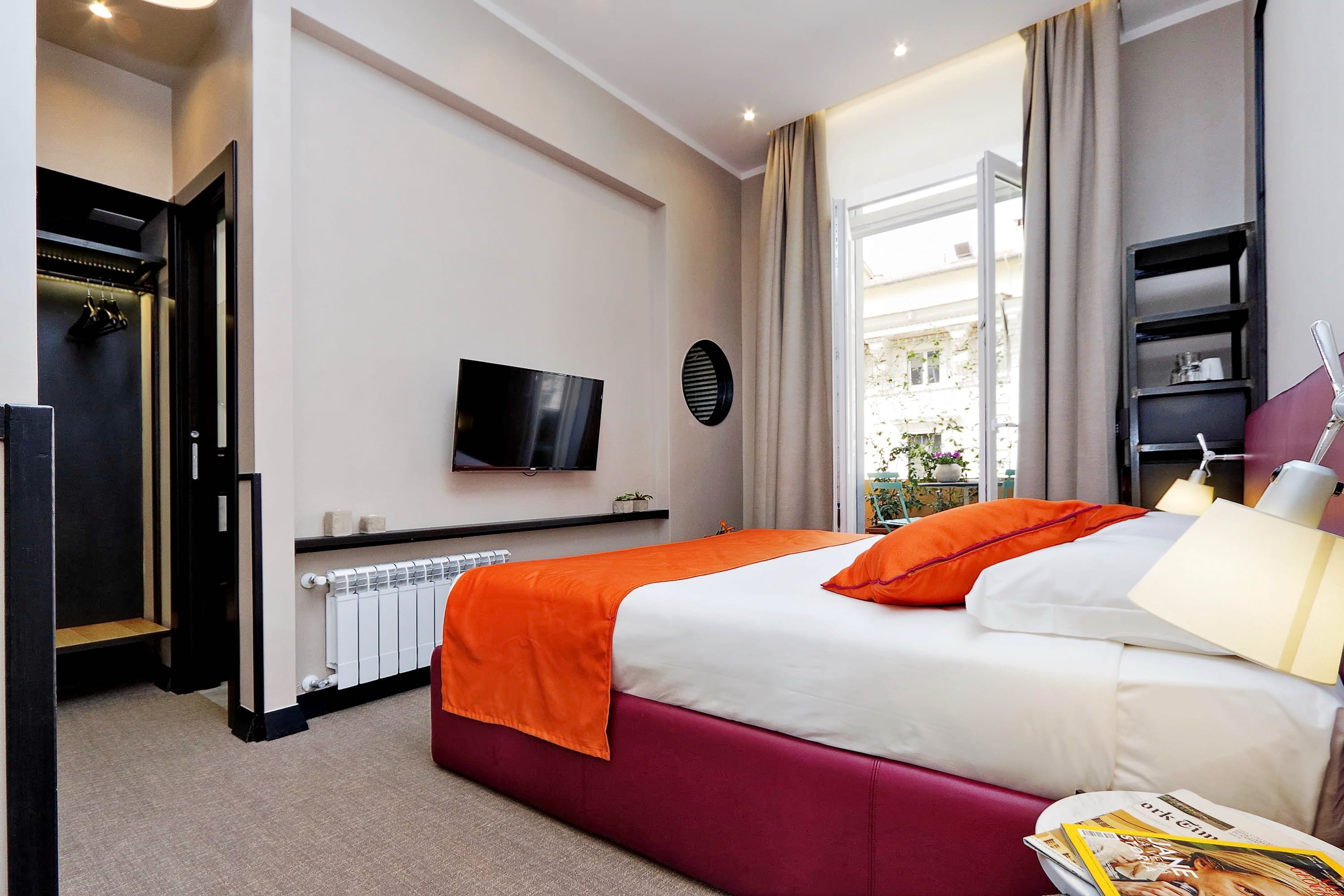 A hotel room with a bed, a TV, a radiator, a window with curtains, and a bookshelf.