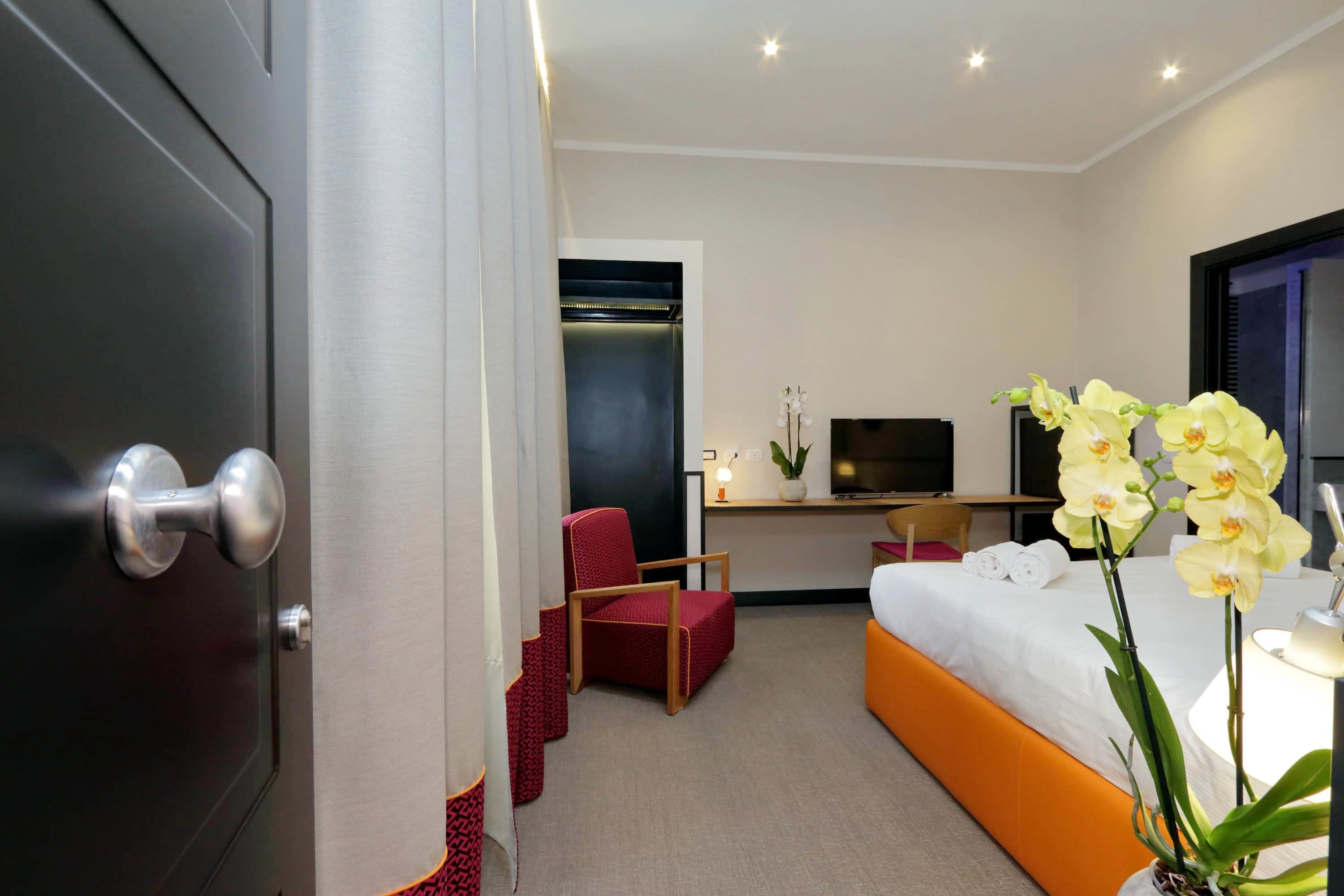 A hotel room with a bed, a desk with a TV, a red chair, and a potted orchid.