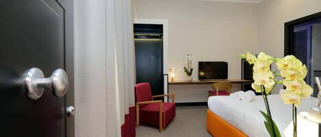 A hotel room with a bed, a desk with a TV, a red chair, and a potted orchid.