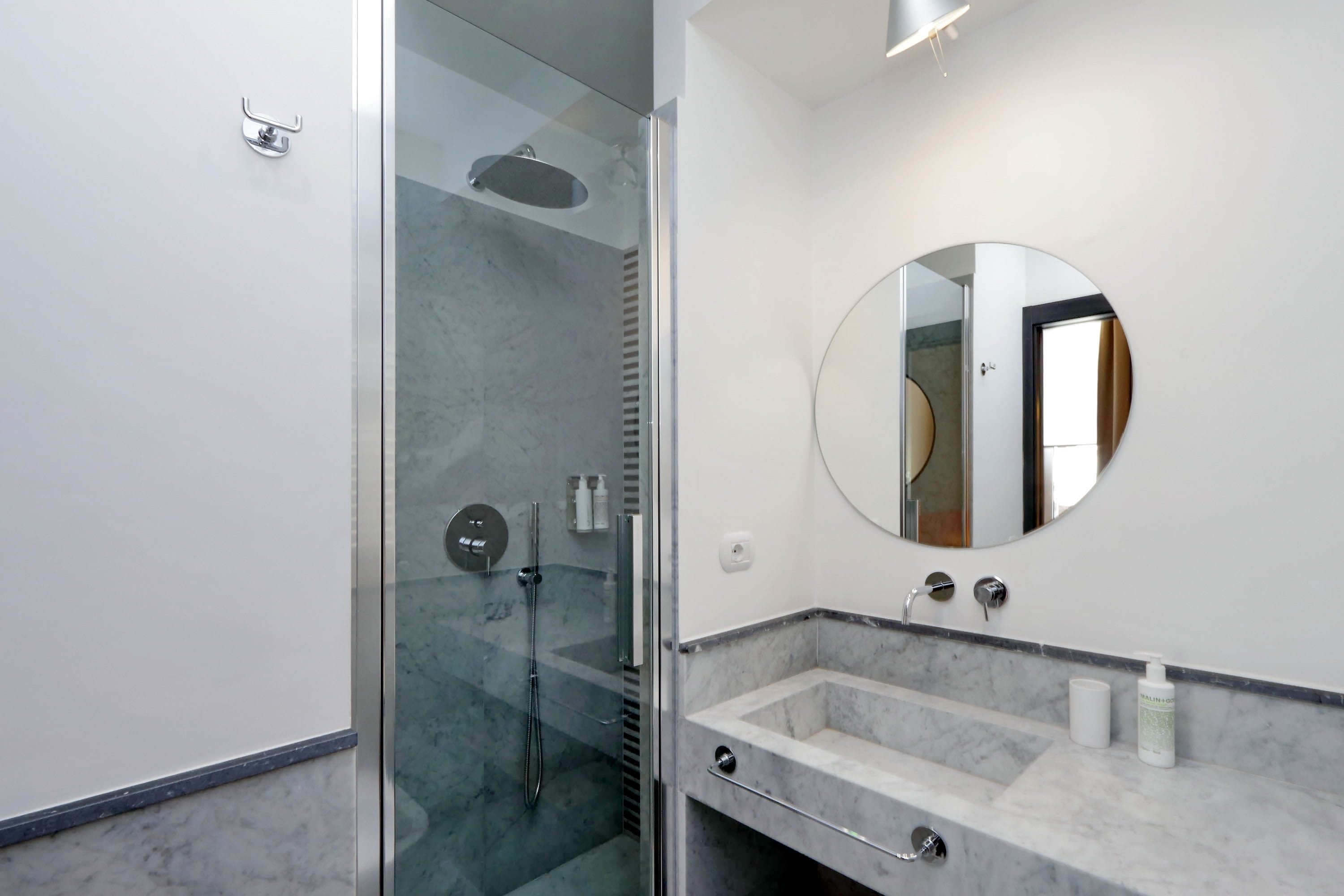 Ein modernes Badezimmer mit einer in Glas eingefassten Dusche, einem мраморной Waschtisch und einem runden Spiegel.