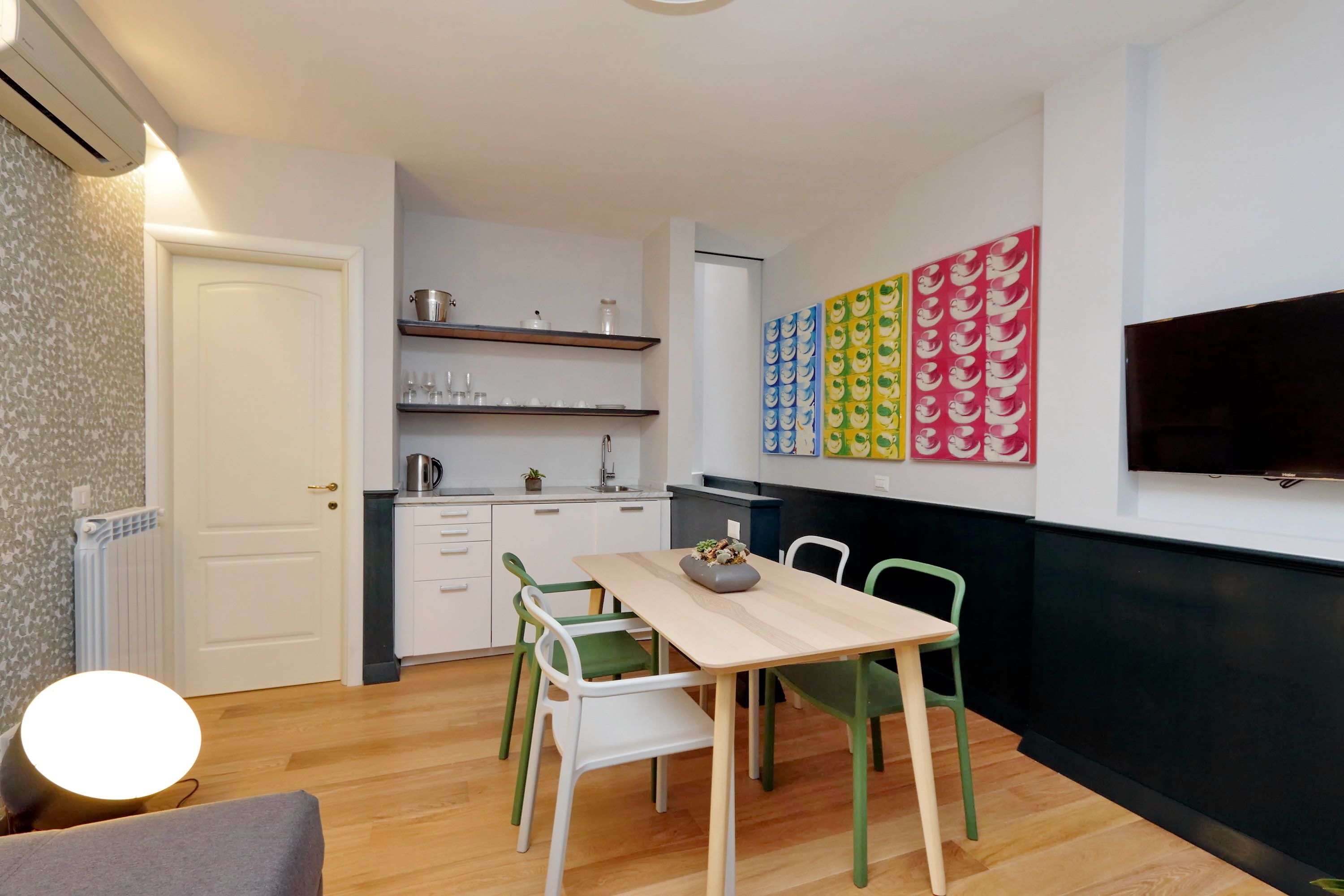 A modern kitchen with white cabinets, a wooden dining table, and green chairs. There is a flat-screen TV mounted on the wall and a colorful abstract art piece above the counter.