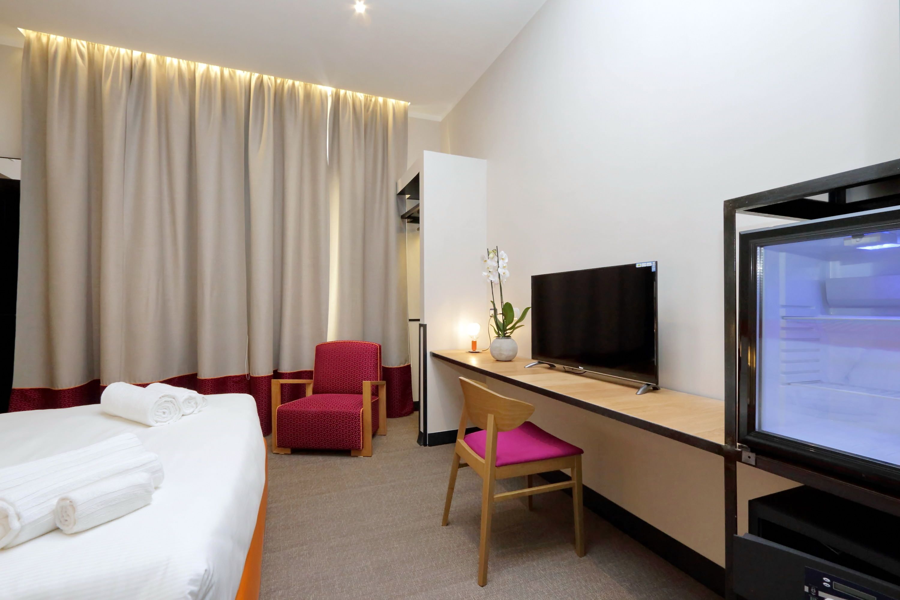 A hotel room with a bed, a desk with a TV, a chair, and a desk lamp.