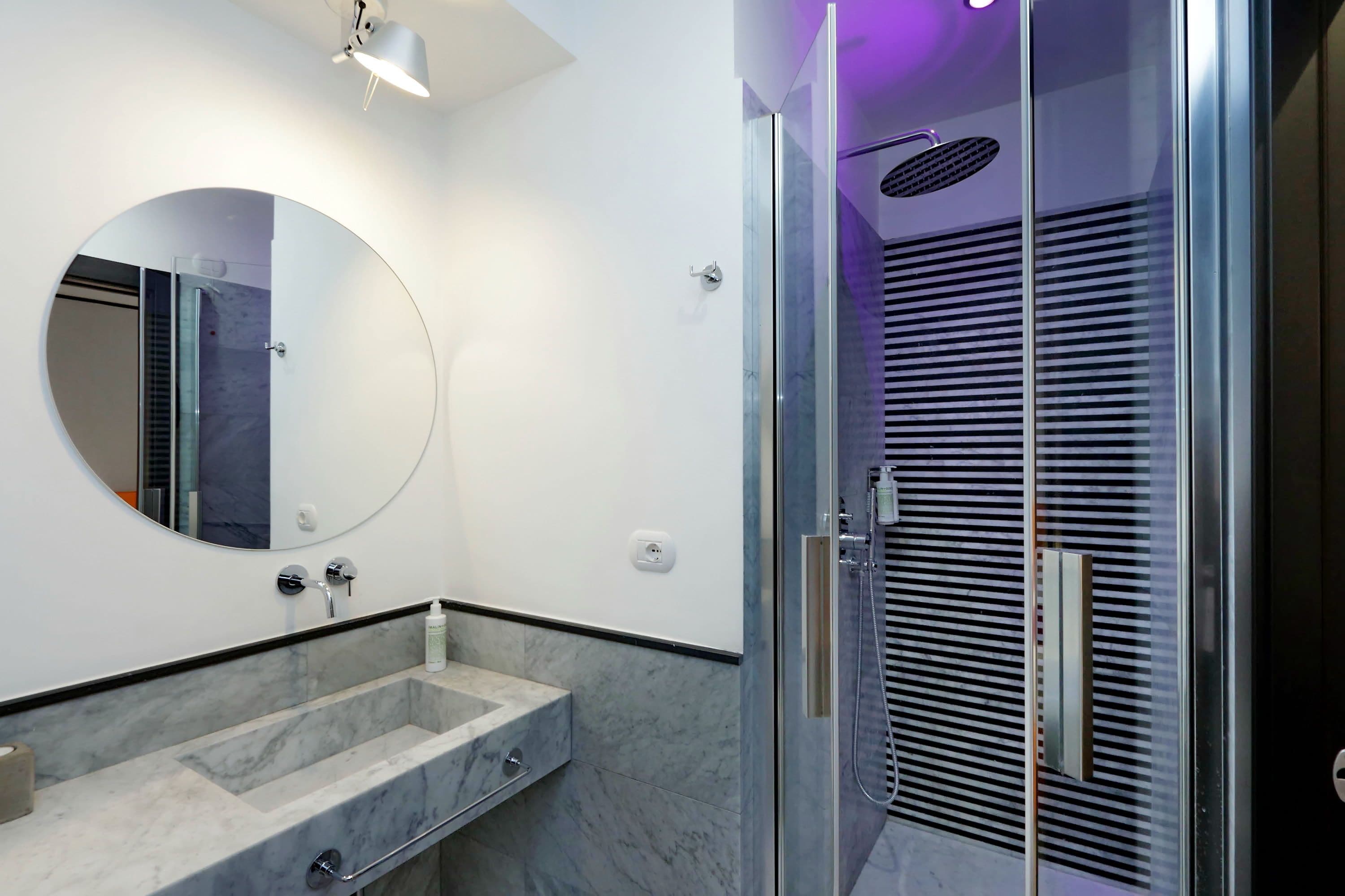 Un baño moderno con un espejo redondo, un lavabo de mármol y una ducha con cerramiento de vidrio y sistema de ventilación.