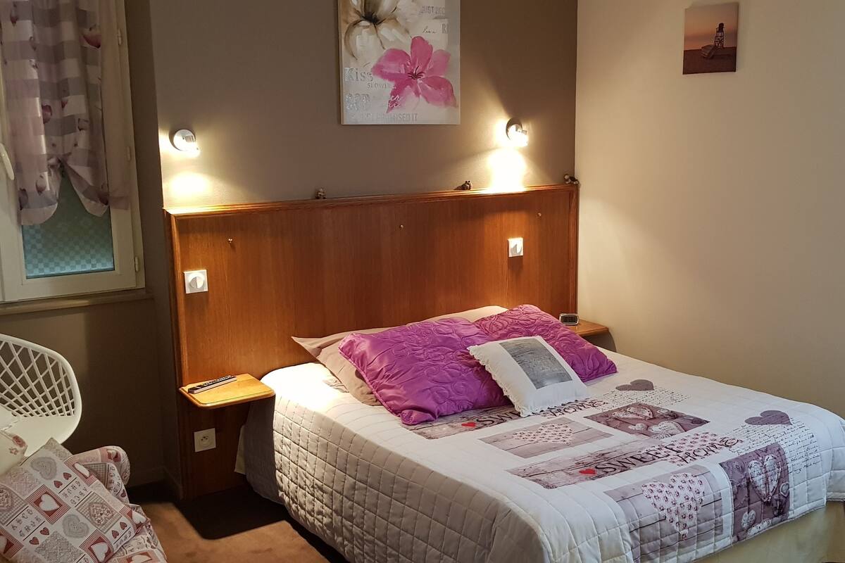 A bedroom with a wooden headboard, a bed with a quilted bedspread and multiple pillows, a bedside table with a lamp, and a chair with a cushion.