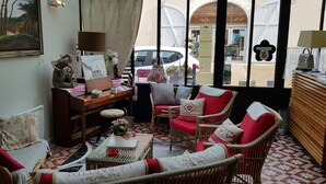 A cozy room with wicker furniture, a desk with a lamp, and a view of the street through large windows. - Au Bosquet Fleuri (Martizay)