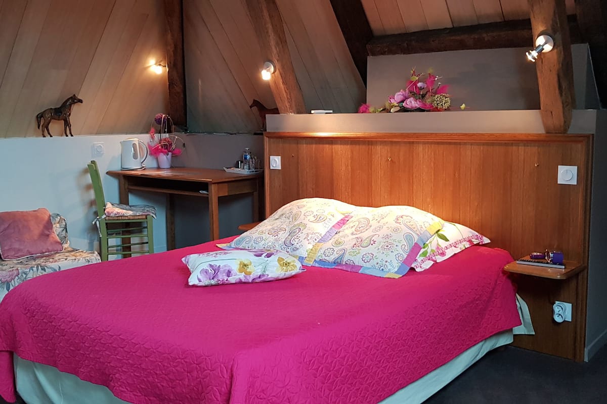 A cozy bedroom with a wooden ceiling, a bed with a pink blanket, a desk with a lamp, and a small shelf with flowers.