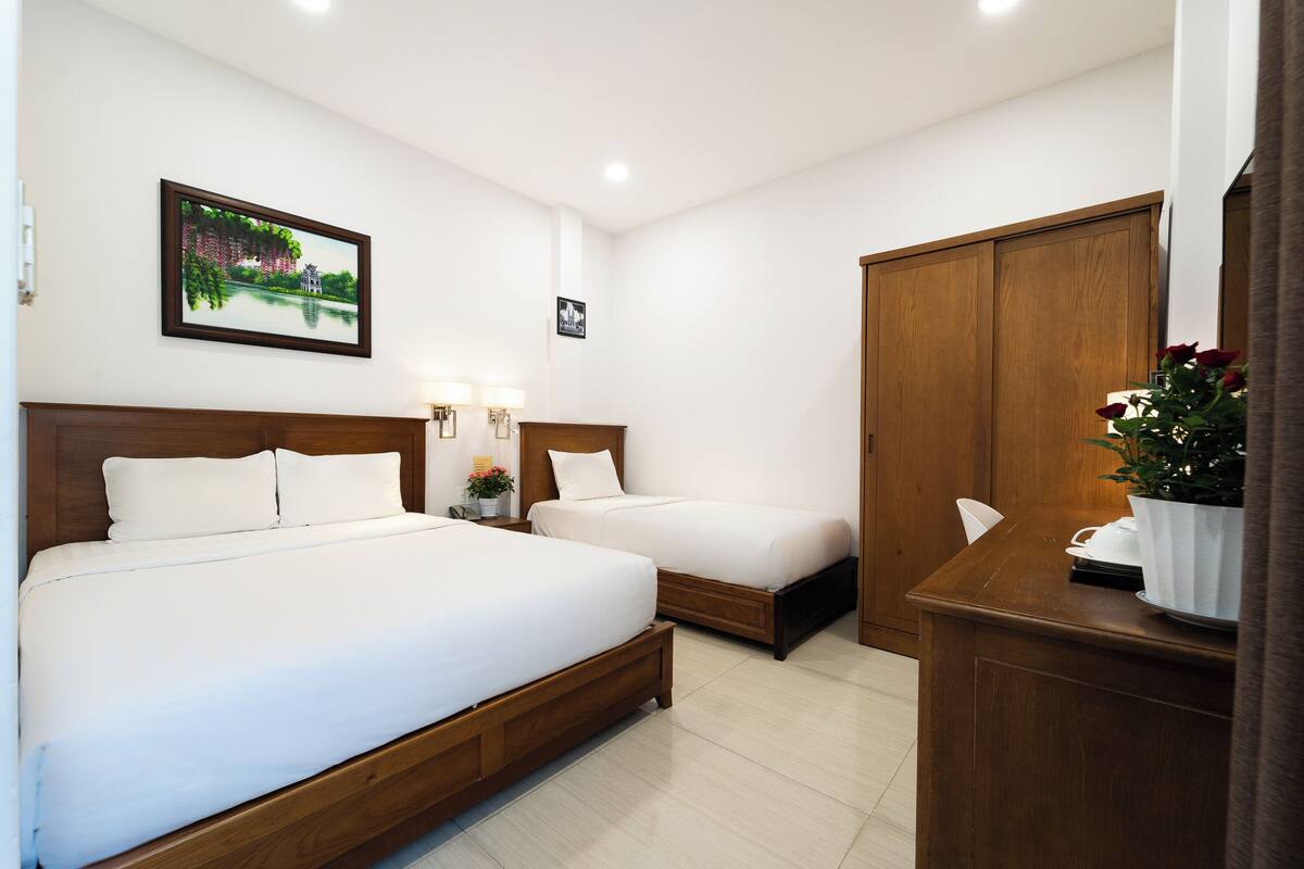 A hotel room with two beds, a wooden wardrobe, a desk with a potted plant, and a painting on the wall.