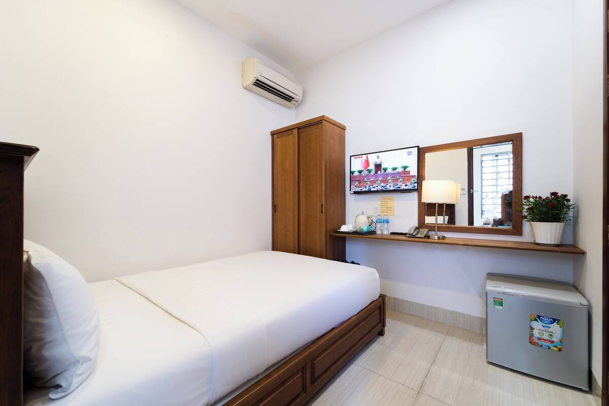 A bedroom with a bed, a wardrobe, a small refrigerator, a mirror, and a shelf with toiletries.