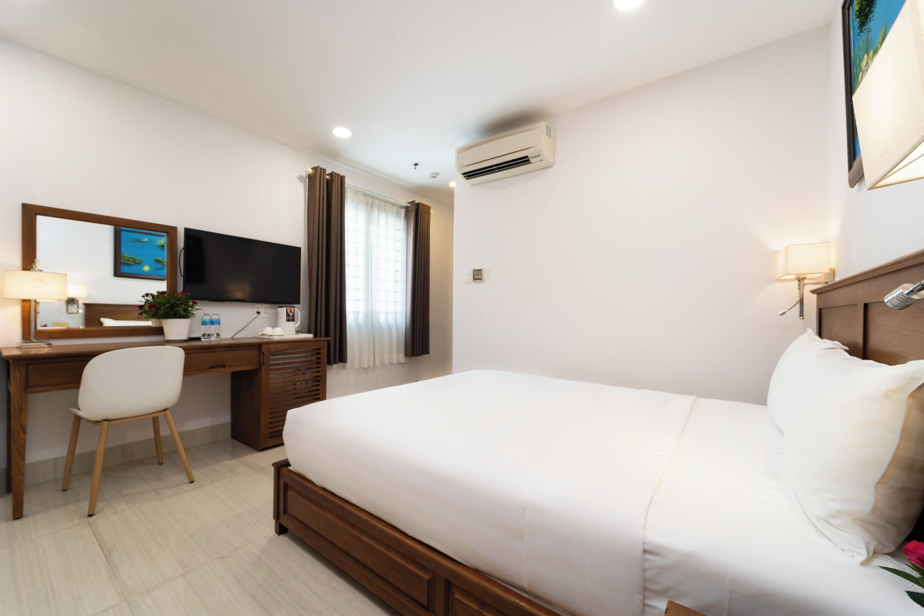 A hotel room with a bed, a desk with a chair, a flat-screen TV, an air conditioner, and a window with curtains.
