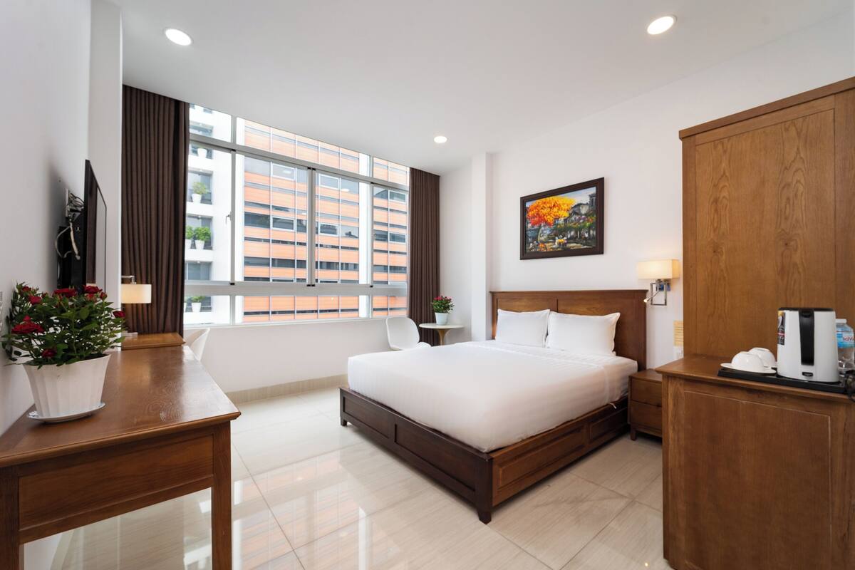 A hotel room with a large bed, a desk with a plant, a small table with a lamp, a wardrobe, and a painting on the wall.
