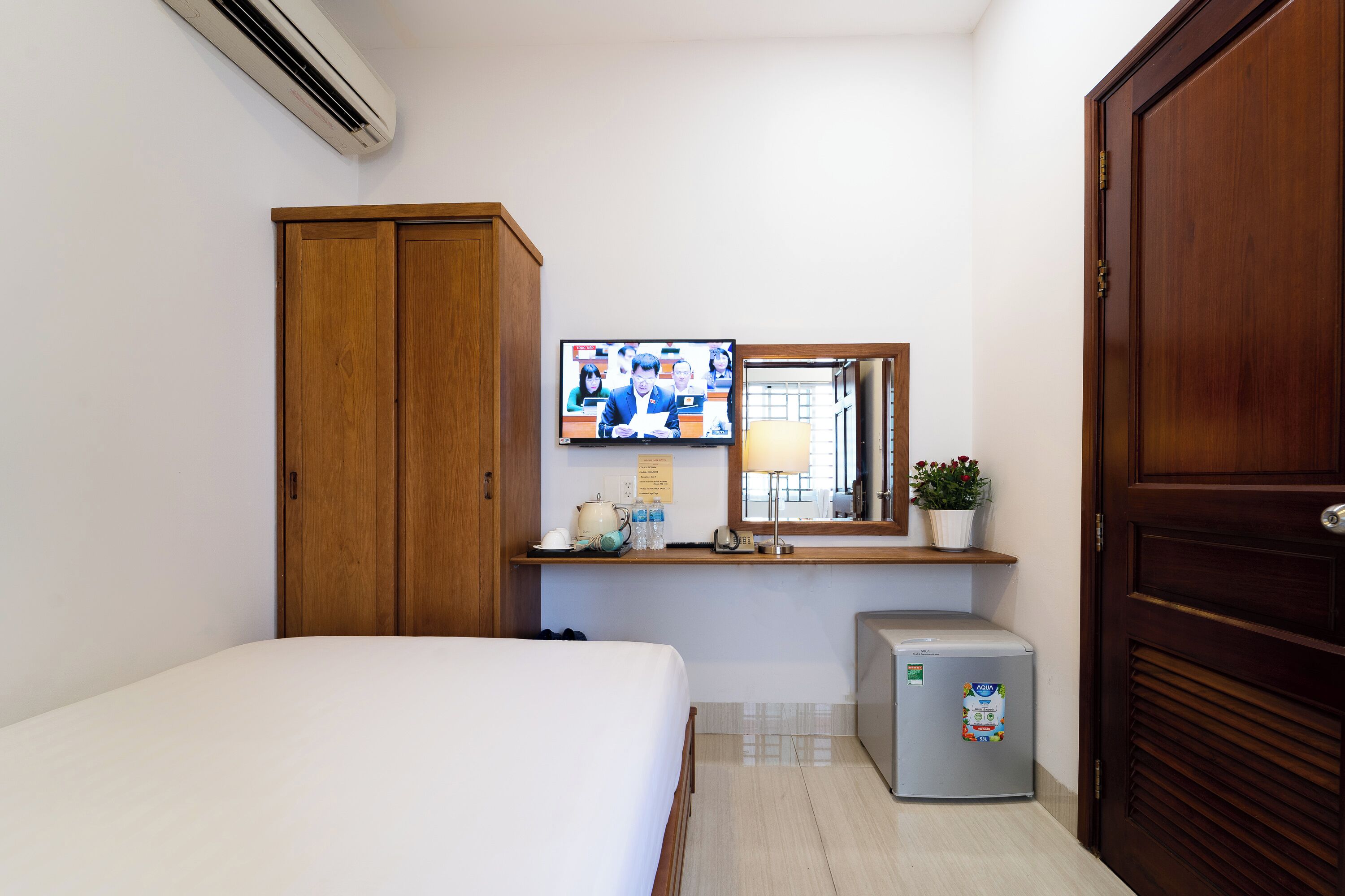 A hotel room with a bed, a small refrigerator, a wooden wardrobe, a television mounted on the wall, and a potted plant on the windowsill.