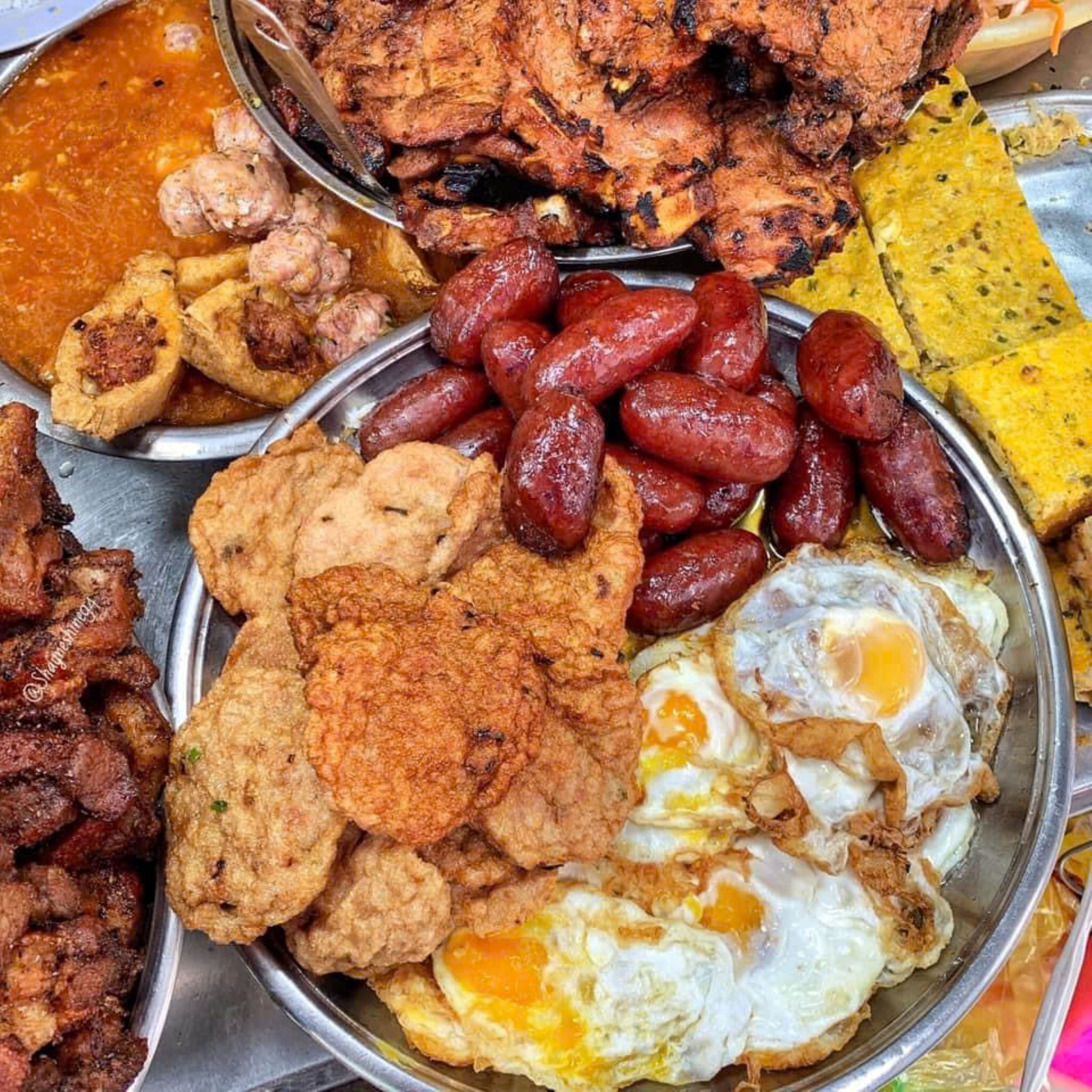 A variety of food items including grilled meat, sausages, fried eggs, and other dishes on a metal platter.