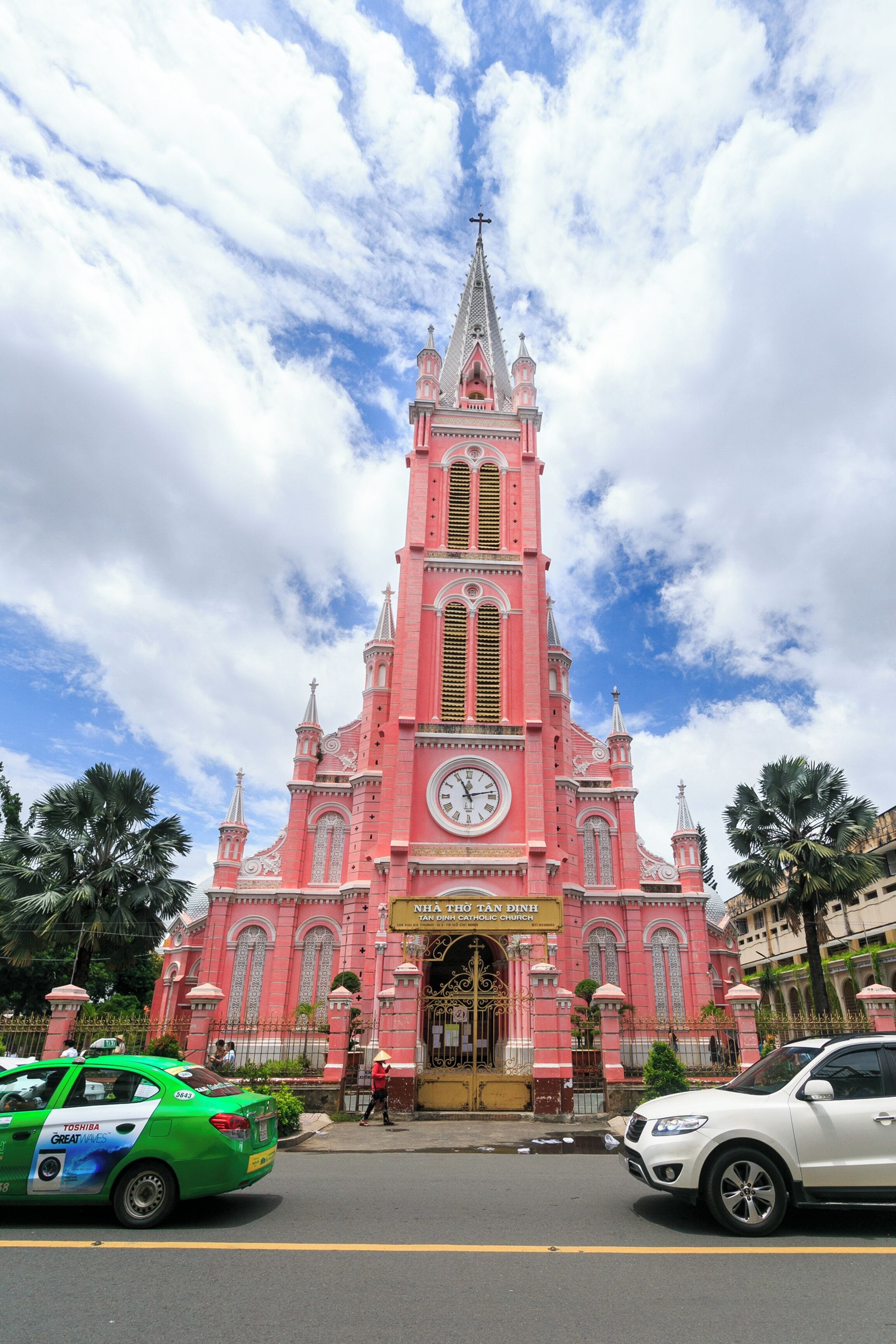 A pink church with a clock tower, surrounded by palm trees and cars on the street.