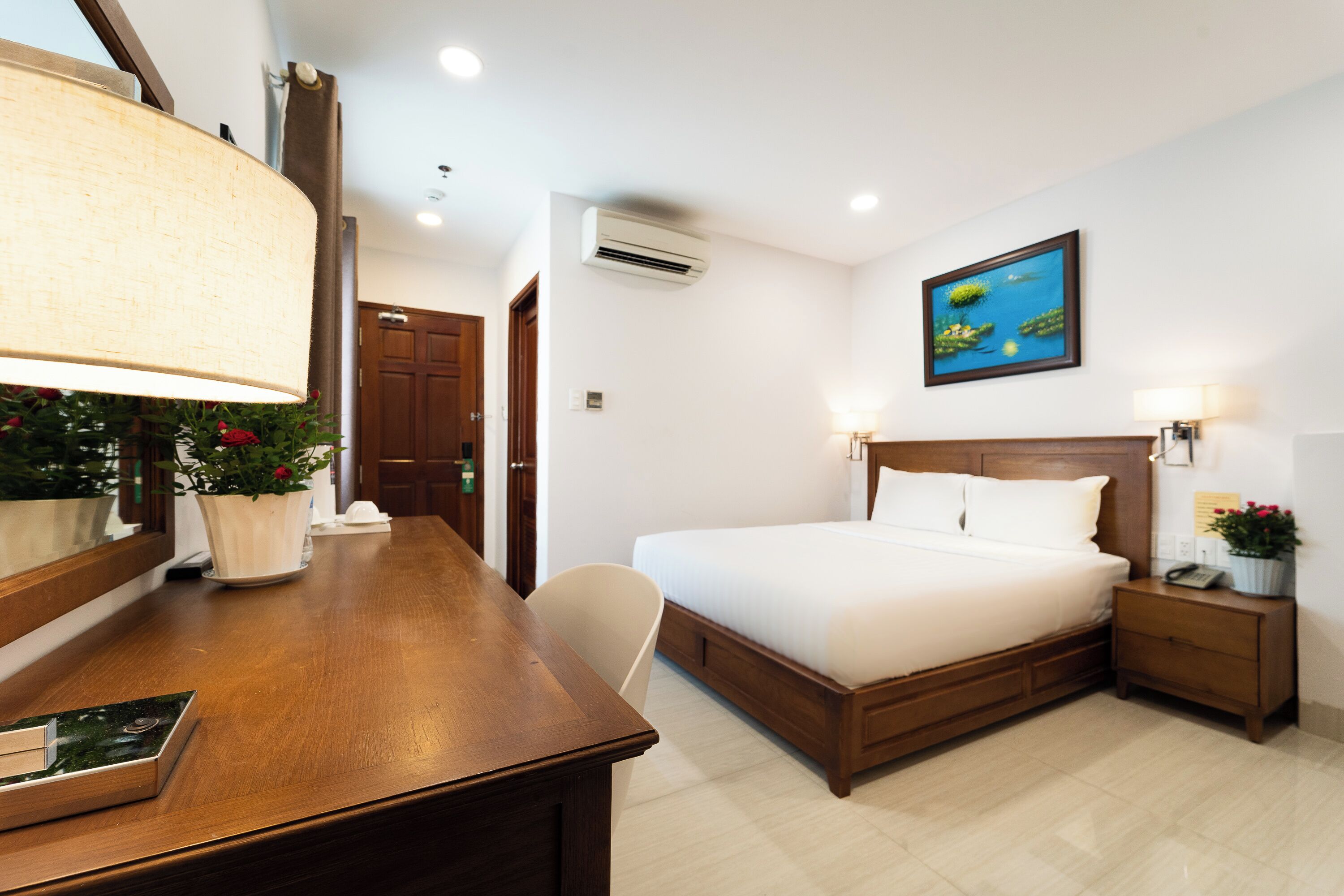 Habitación de hotel con cama de madera, un escritorio con lámpara, una silla, mesita de noche y aire acondicionado instalado en la pared.