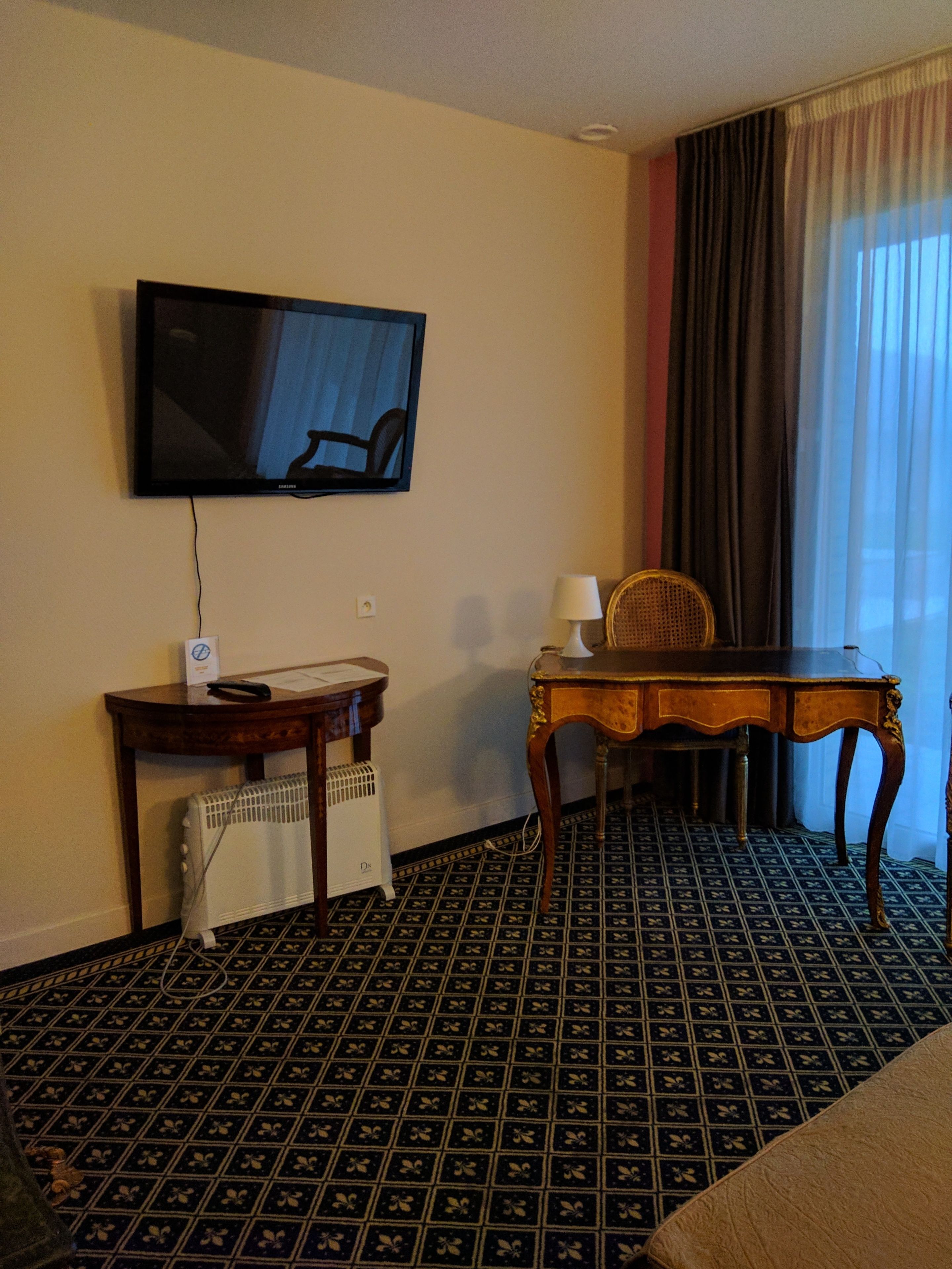 Habitación de hotel con un televisor montado en la pared, una mesita con una silla, un radiador y una alfombra estampada.