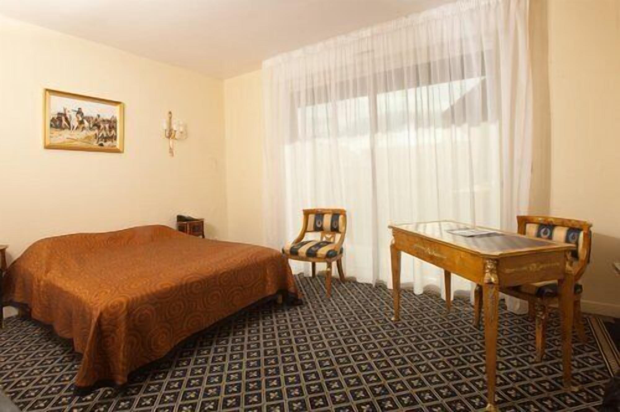 A hotel room with a large bed, a wooden table, two chairs, a painting on the wall, and a patterned floor.