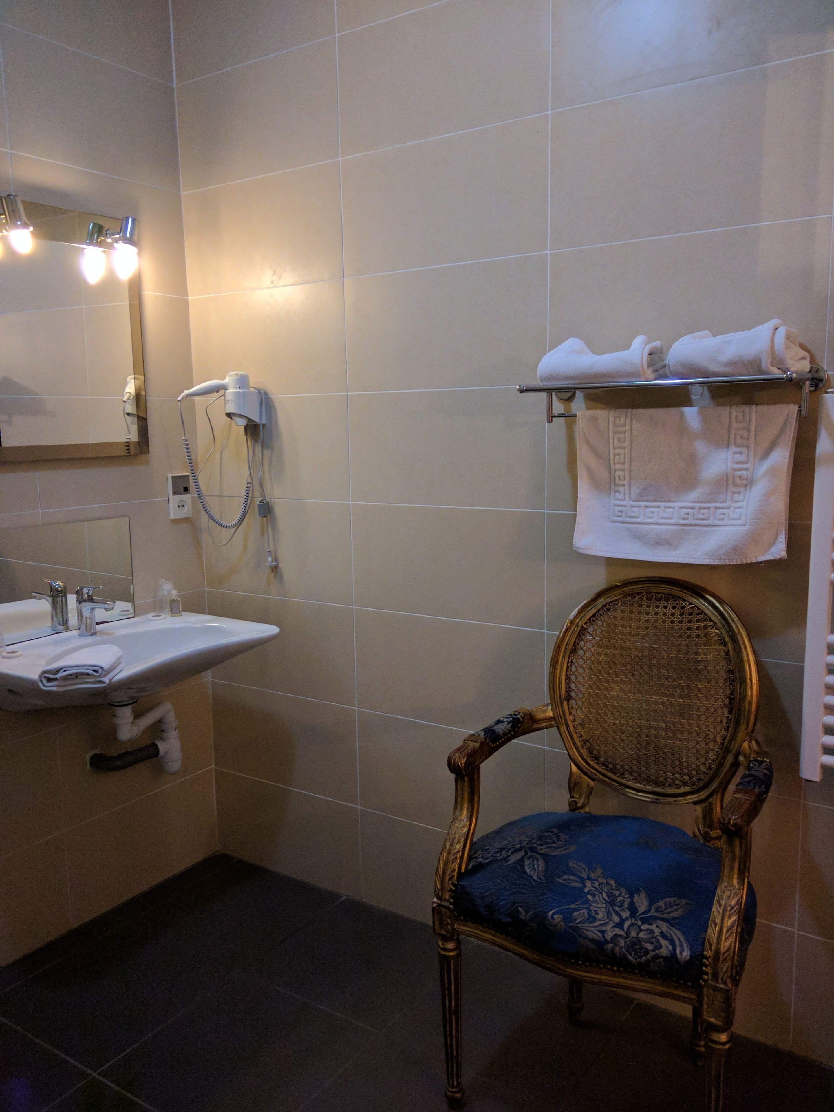 Een badkamer met een wastafel, spiegel, haardroger, handdoekhouder en een stoel.