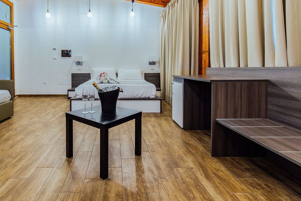 Quarto de hotel moderno com piso de madeira, cama com roupa de cama branca, uma pequena mesa com um vaso e um banco de madeira escura.