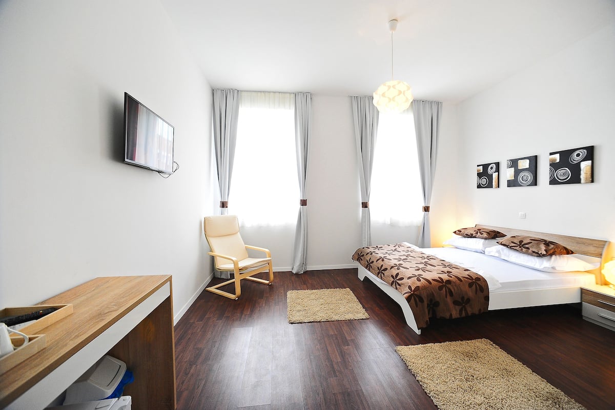 A modern bedroom with a wooden desk, a comfortable chair, a bed with a floral bedspread, and a TV mounted on the wall.