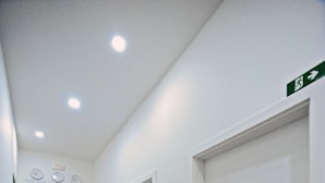 Un couloir aux murs blancs, doté d’un plancher en bois et de portes numérotées. On y trouve des horloges murales et une petite étagère sur laquelle sont posés divers objets.