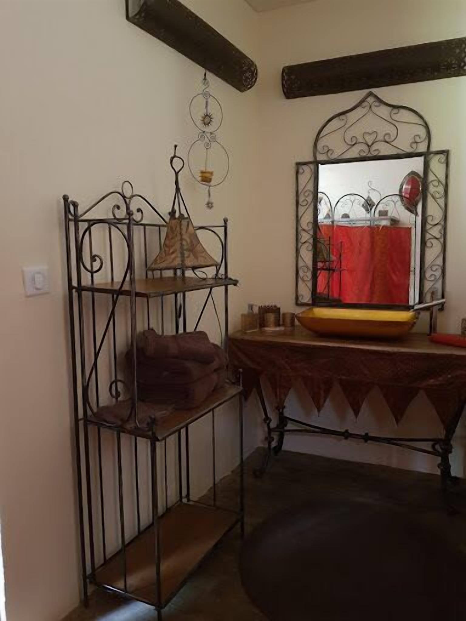 A room with a metal-framed shelf unit holding a lamp, a mirror with a decorative frame, and a table with a bowl and a red cloth.