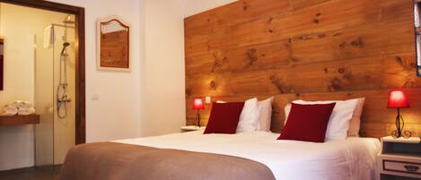 Habitación de hotel con una cama grande, cabecera de madera, dos mesitas de noche con lámparas, panel de pared de madera, zona de ducha y un perchero para toallas.