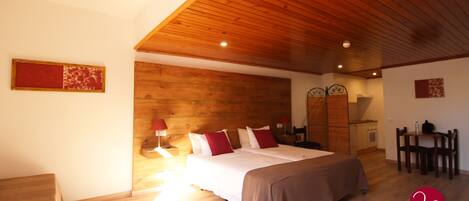 Quarto com teto de madeira, cama com roupa de cama vermelha e branca, mesa de cabeceira com abajur e uma pequena cozinha americana no canto.