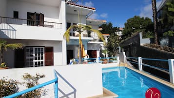 A swimming pool adjacent to a white building with balconies, a palm tree, and a clear blue sky.